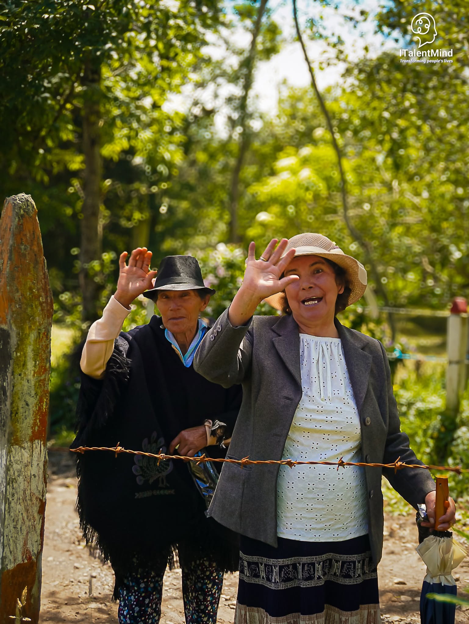 Dos personas mayores con sombrero saludan sonrientes en un espacio al aire libre.